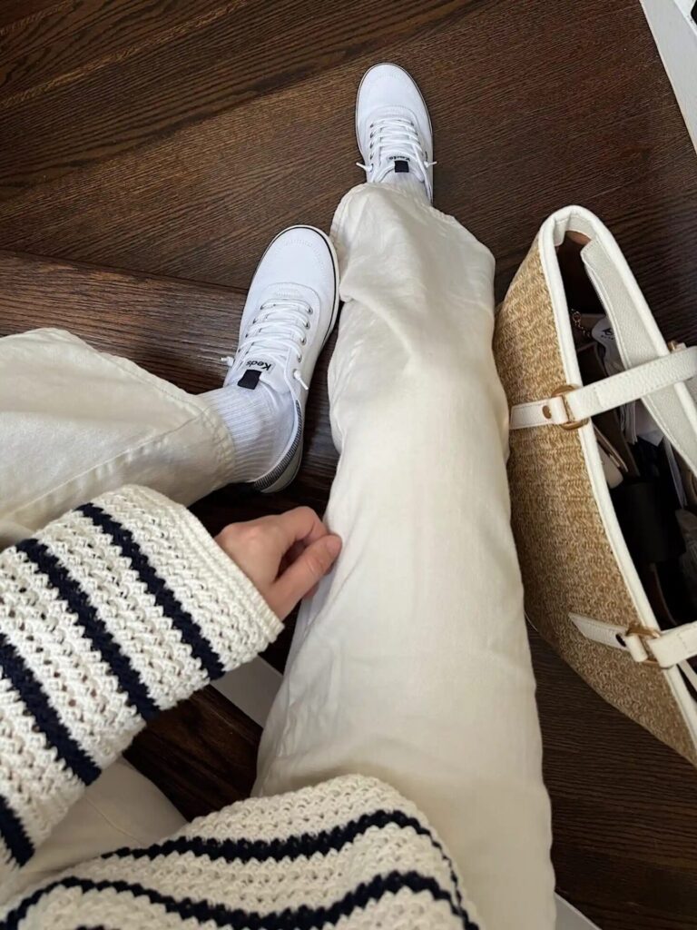 Person wearing classic white low-top sneakers with white denim and a striped knit sweater, holding a woven tote.