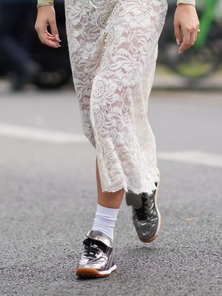 Ultra-realistic photo of a woman wearing a lace maxi skirt paired with metallic silver sneakers on a city street at dusk.