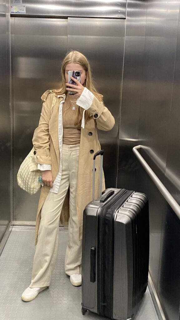 Traveler wearing a beige trench coat with blue jeans and tote bag, walking through terminal