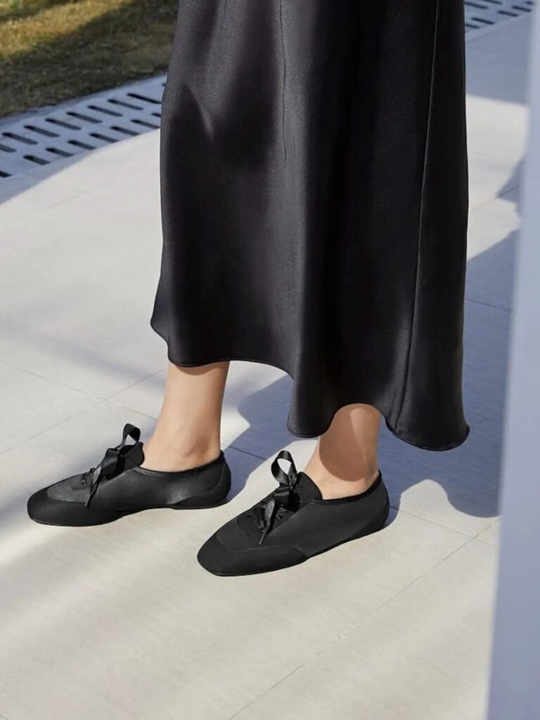Street-style close-up of a person in a black satin skirt and slim retro sneakers with ribbon laces.