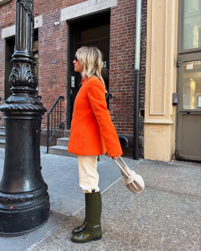 Vibrant fall outfit with a bright orange sweater styled with white pants and olive boots, inspired by the showgirl orange trend of 2025.