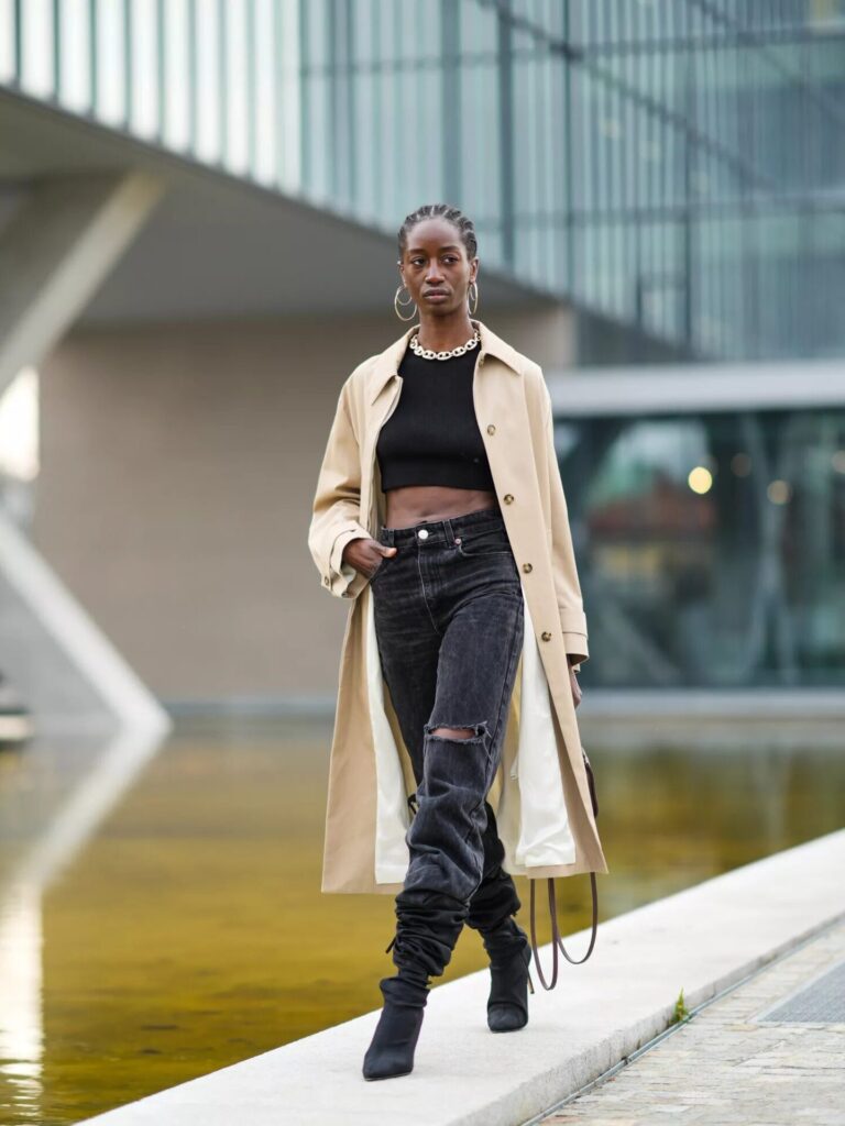 Woman wearing beige trench coat over black ripped jeans, black halter crop top, gold jewelry, and Telfar handbag with ankle boots.