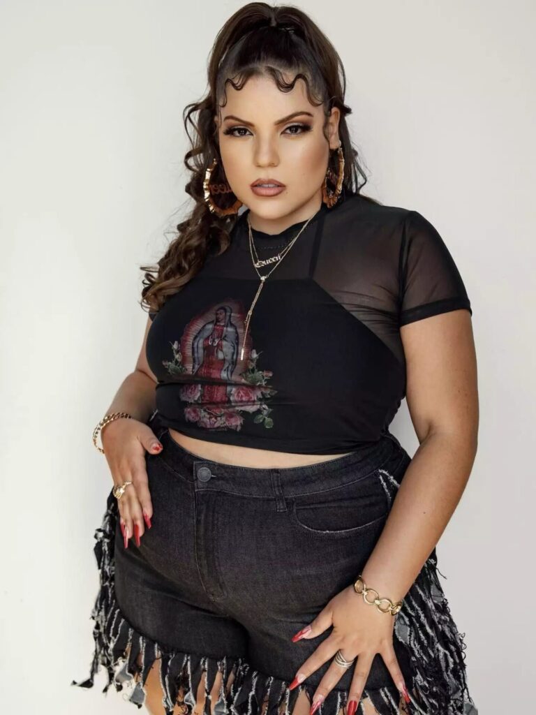 Elle Baez in distressed black denim shorts, sheer lace shirt, graphic cami tank, and oversized hoop earrings styled for a concert.