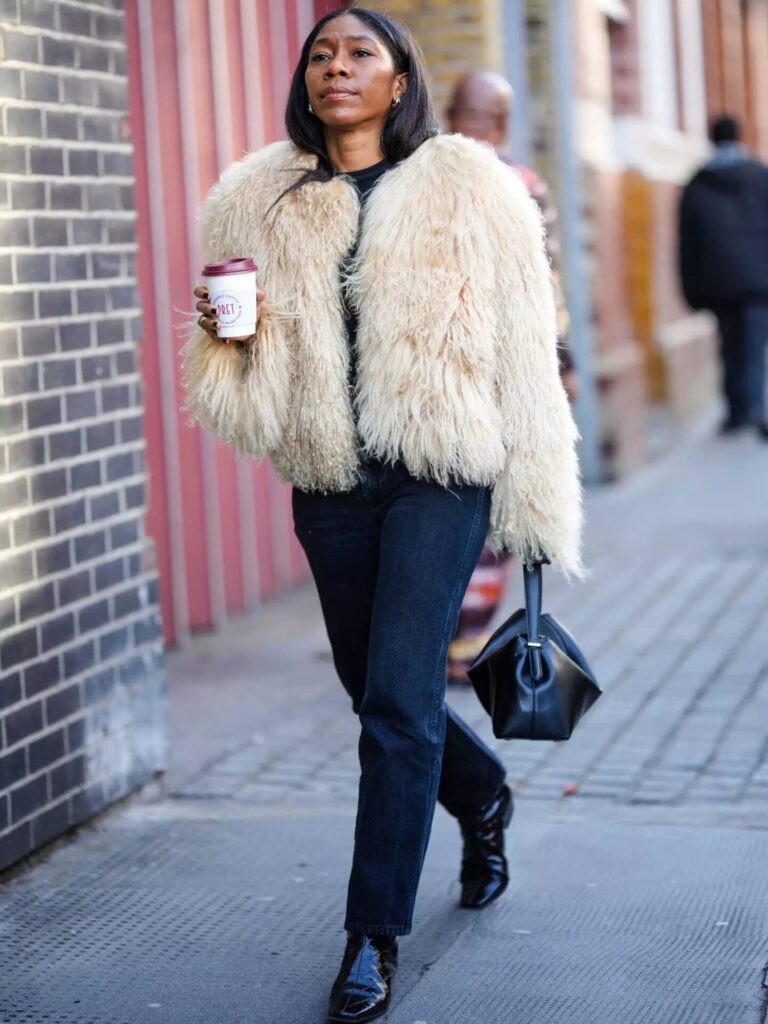 Street style woman in black jeans with a fluffy sherpa jacket, leather handbag, and ankle boots for a cozy chic black denim look.