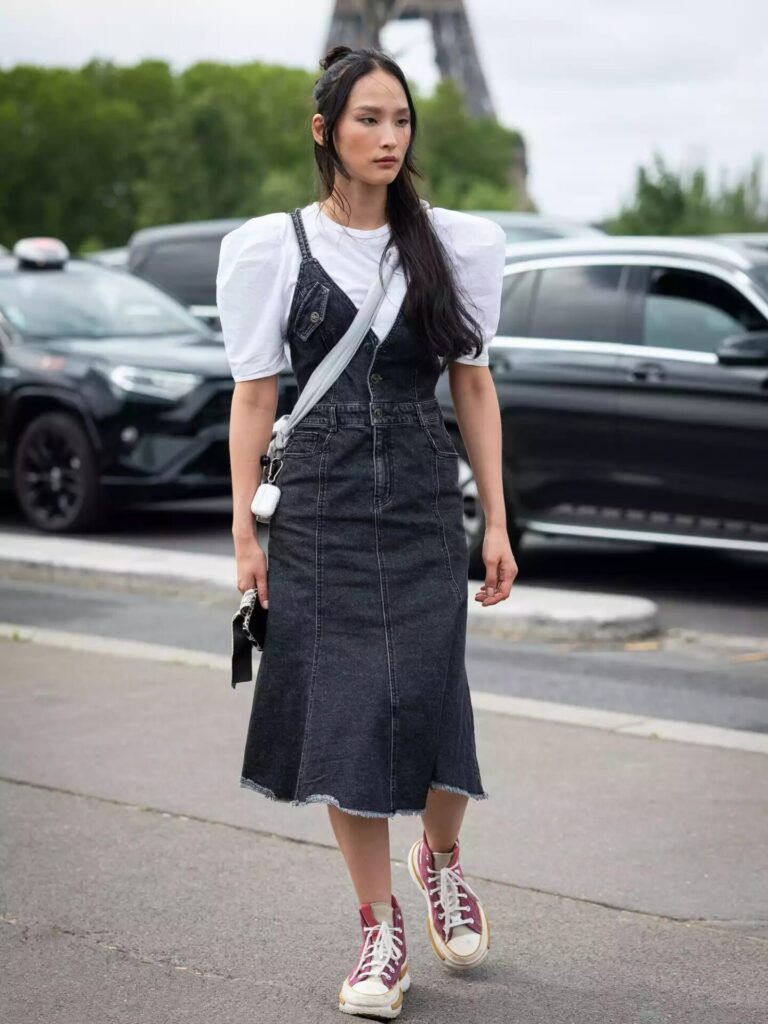 Woman in a sleeveless black denim midi dress layered over a white puff-sleeved tee with pink Converse sneakers and crossbody bag