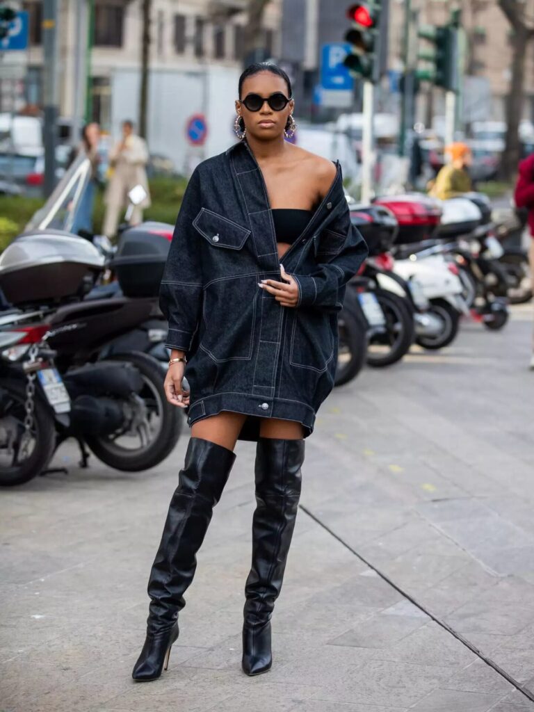 Didi Stone wearing oversized black denim jacket styled as a dress with thigh-high boots, bandeau, and round sunglasses for a bold outfit.