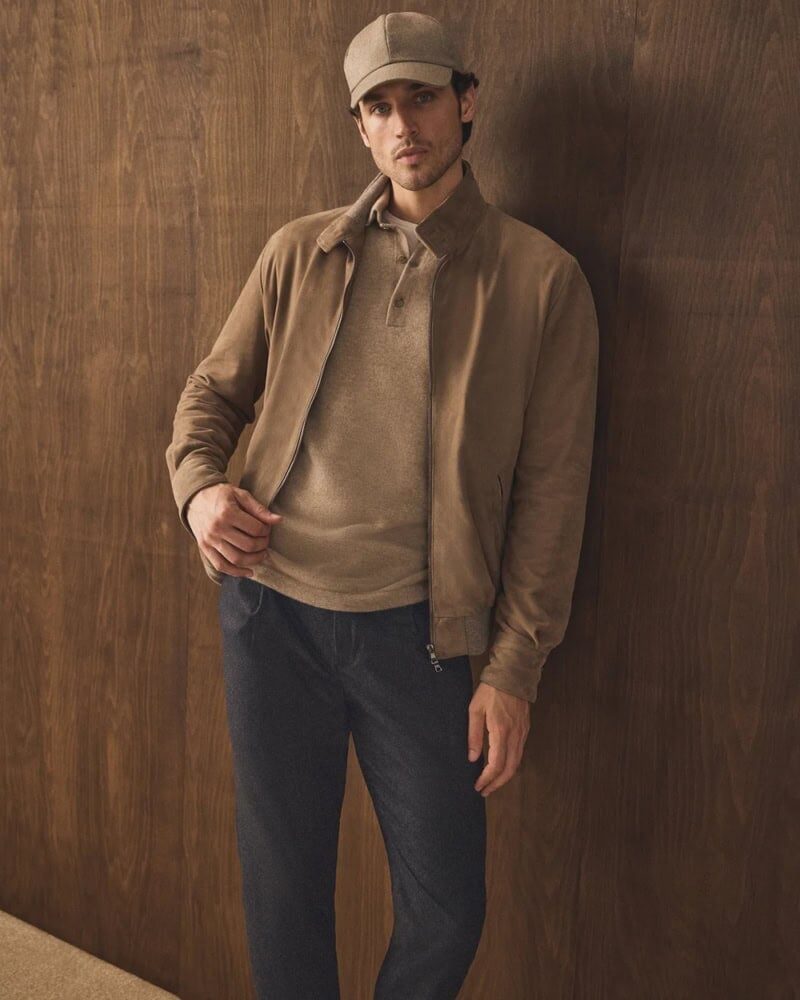 man wearing brown suede bomber jacket with black jeans leaning against wall