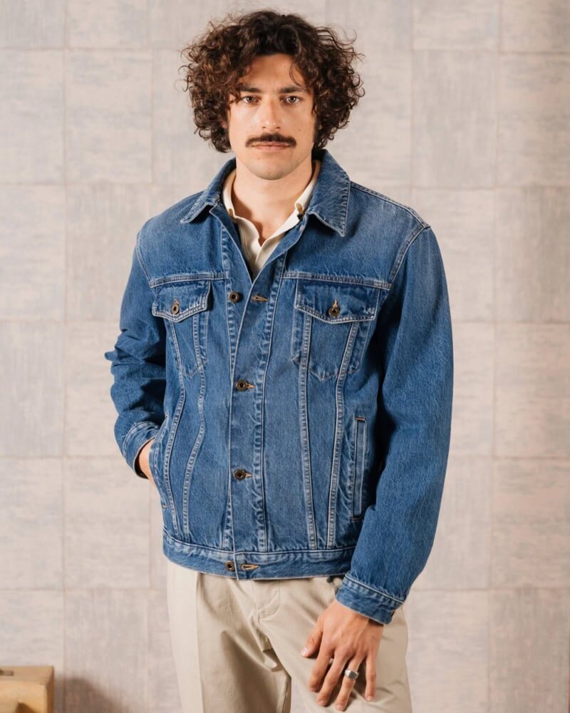man layering blue denim jacket over hoodie and chinos with backpack