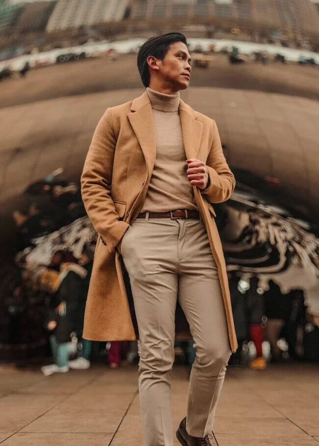 man wearing walnut overcoat and beige turtleneck with brown derby shoes in modern office