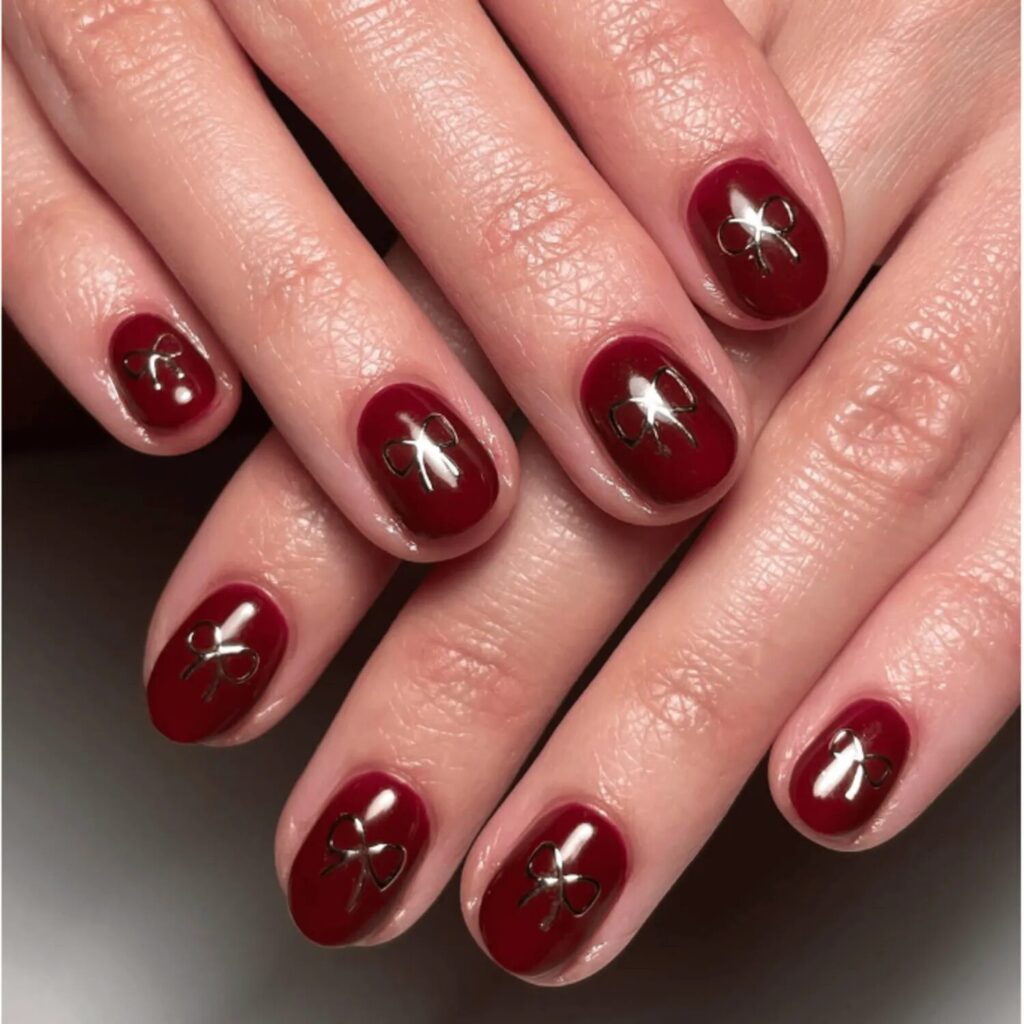 Short nails painted in deep oxblood polish with delicate silver bows and glossy finish