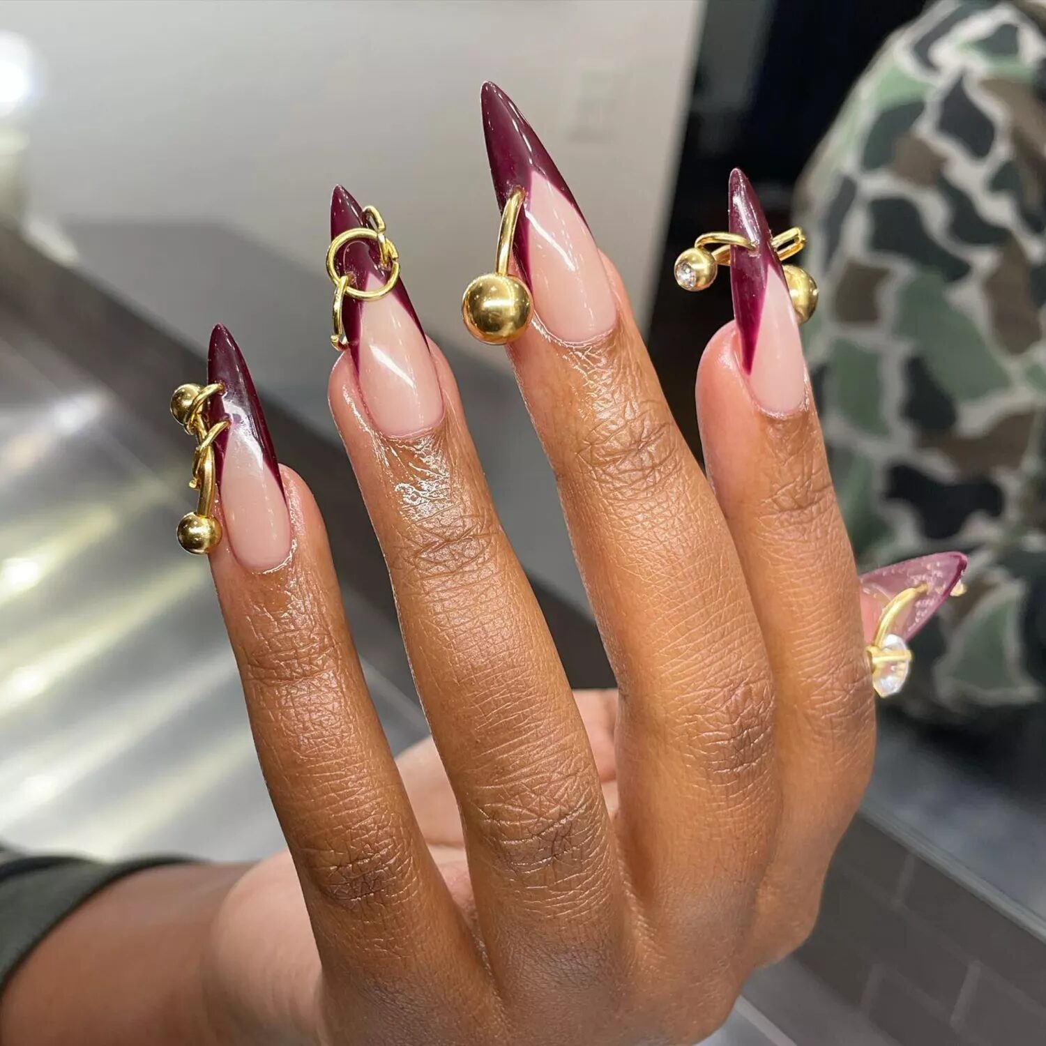 Maroon French tip manicure with gold nail charms and piercings creating a luxurious 3D nail design.