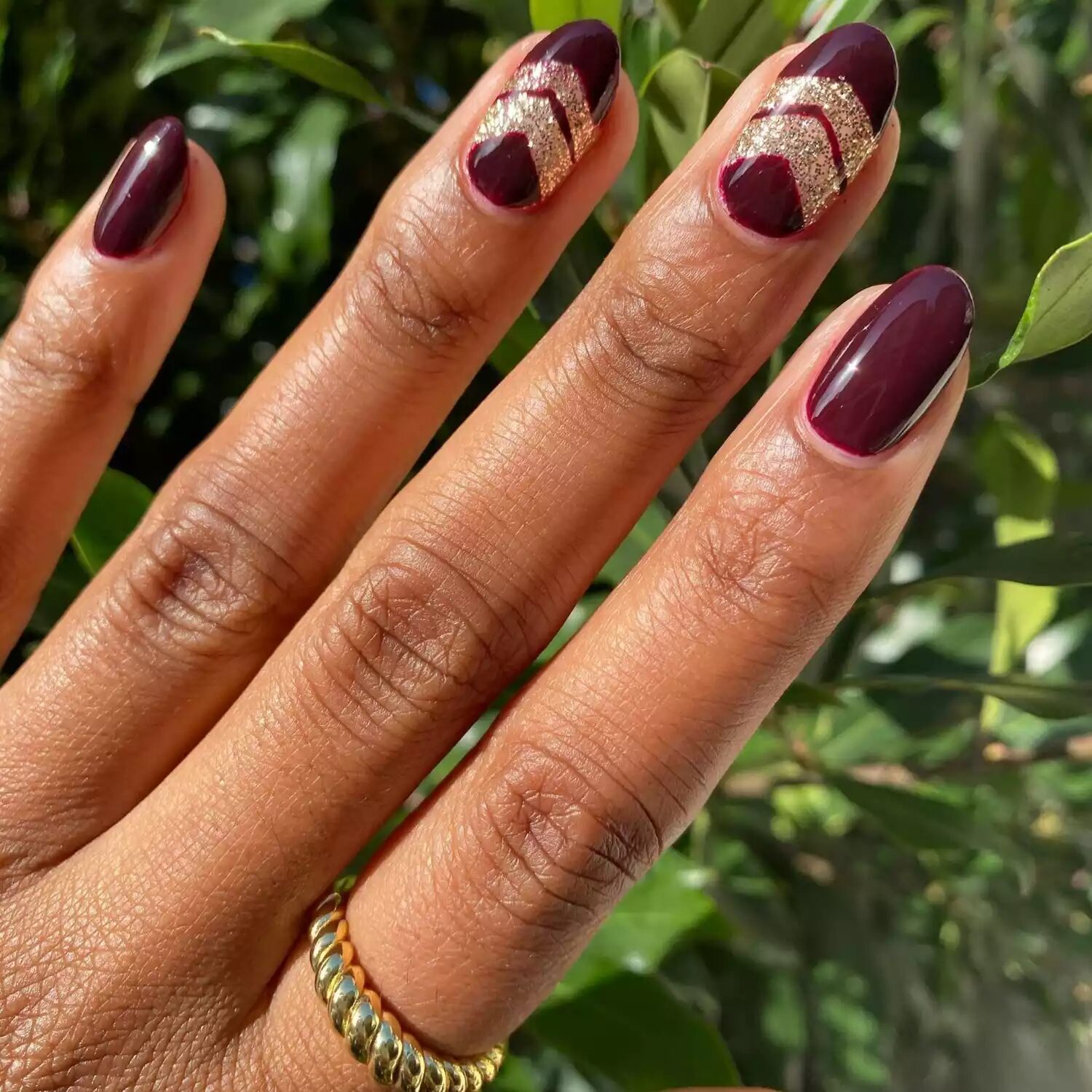 Dark burgundy nails with gold sparkle accents and geometric shimmer shapes for a festive fall manicure.