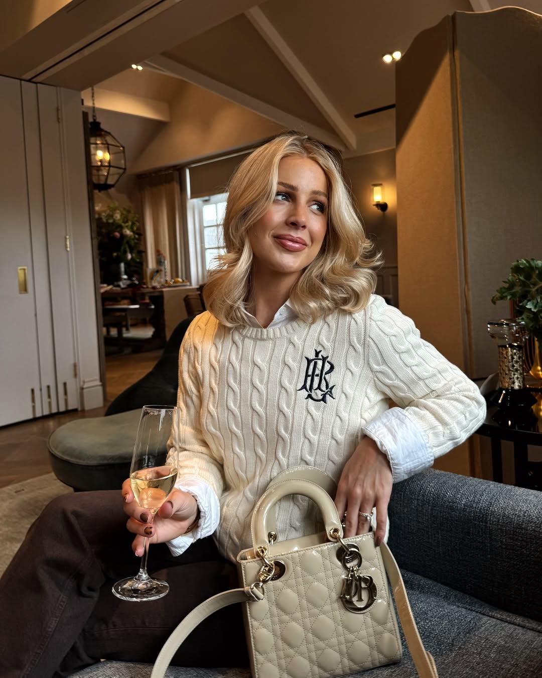Woman wearing a cream cable-knit sweater over a white shirt with dark trousers and a cream bag.