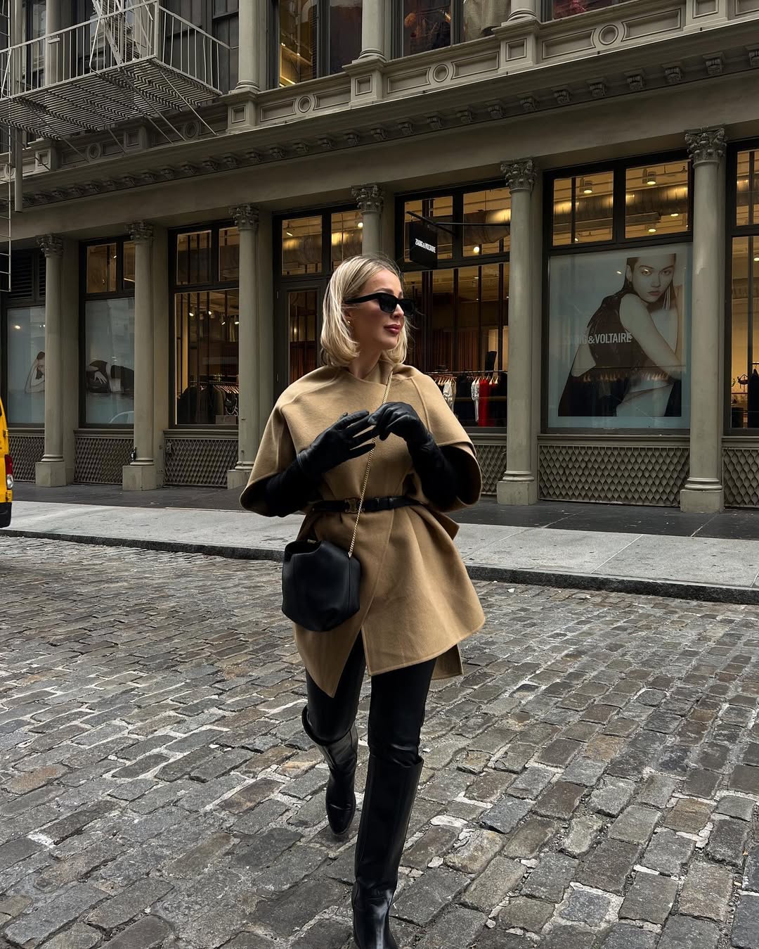 Woman wearing a camel cape coat cinched with a belt and black leather accessories.