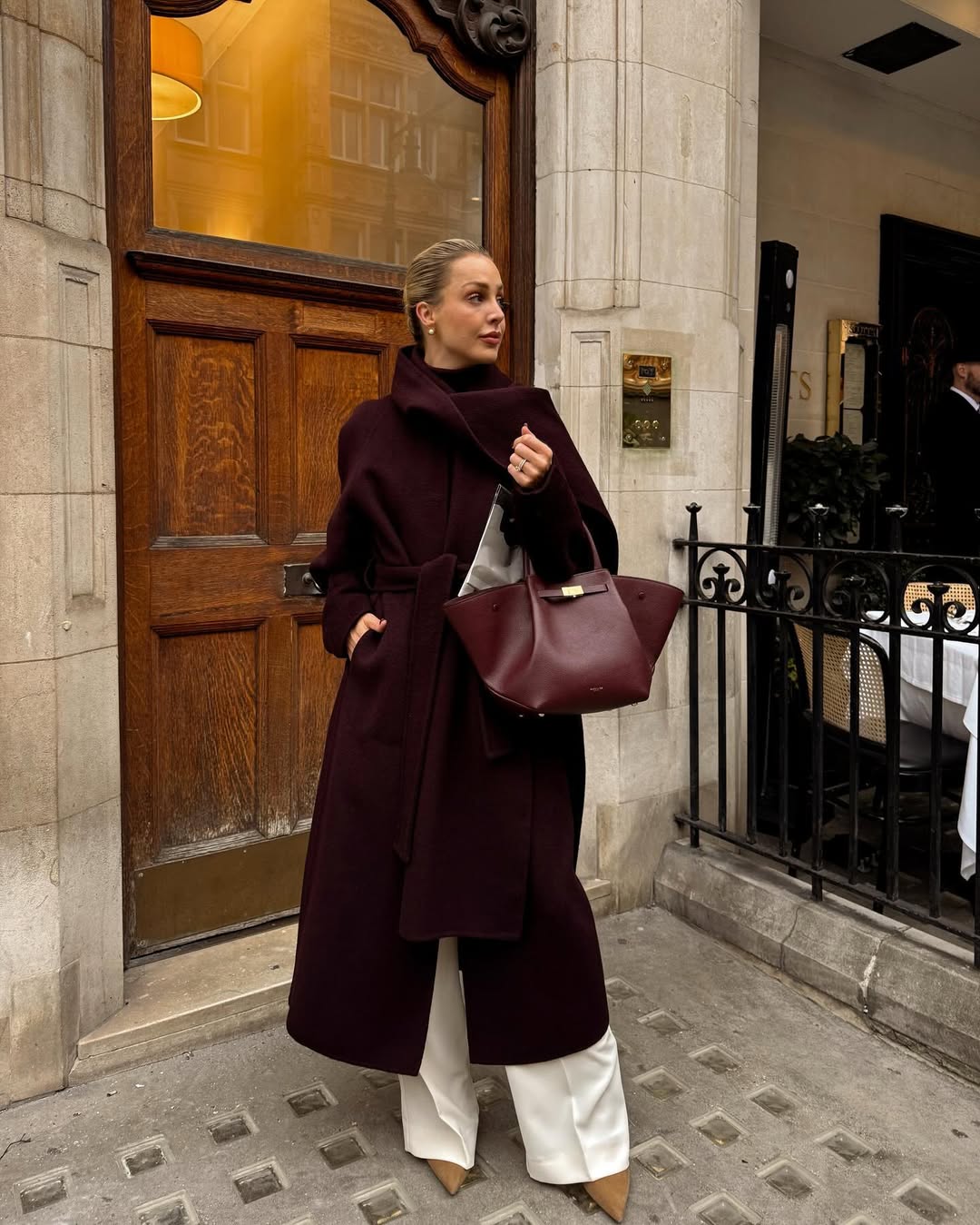 Woman wearing a burgundy wrap coat with cream trousers.