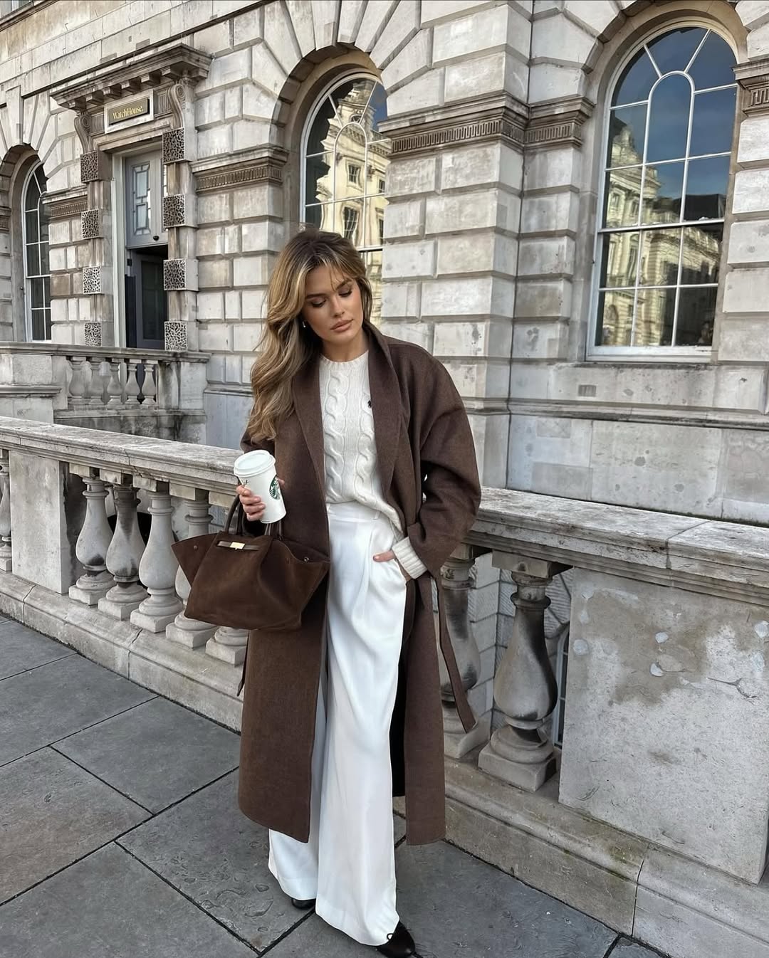 Woman in a brown wrap coat over a cream knit sweater with cream trousers.