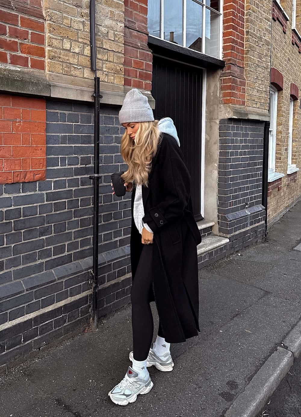 a woman wearing black leggings, a grey hoodie, a black overcoat, chunky sneakers, and a beanie