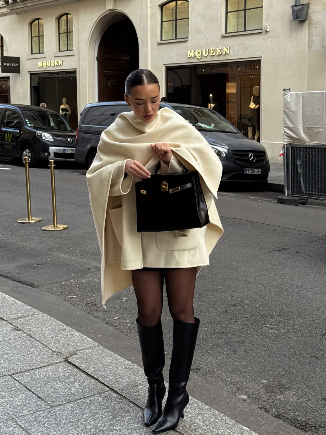 Woman in a cream cape coat with sheer tights and black leather boots.