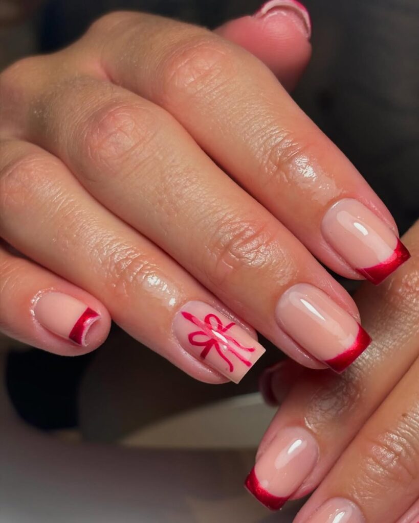 Square nails with bold red French tips and a single accent nail styled as a wrapped gift with a bow.