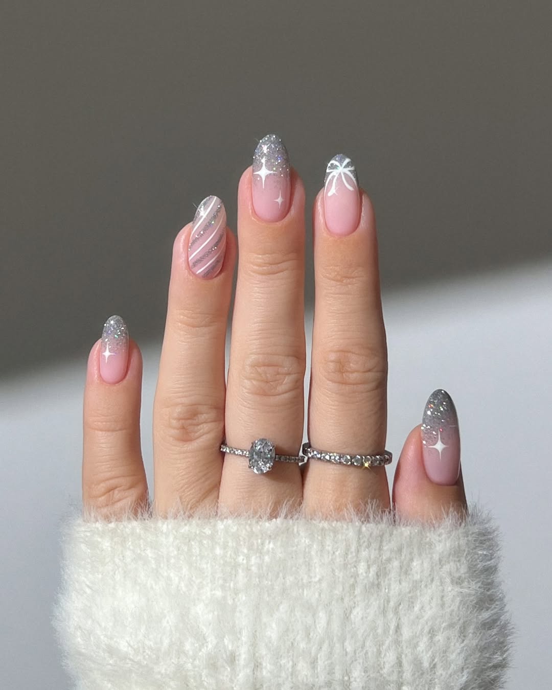 Rosy nude nails with fine silver glitter tips, white starbursts, stripes, and a small bow accent.