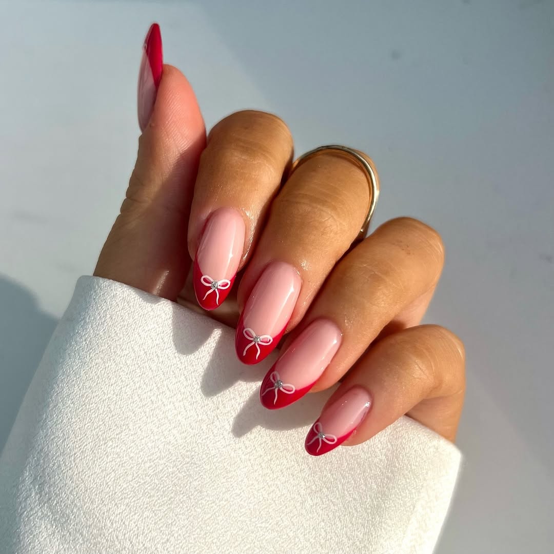 Soft nude nails featuring bold red French tips and small white bow details.