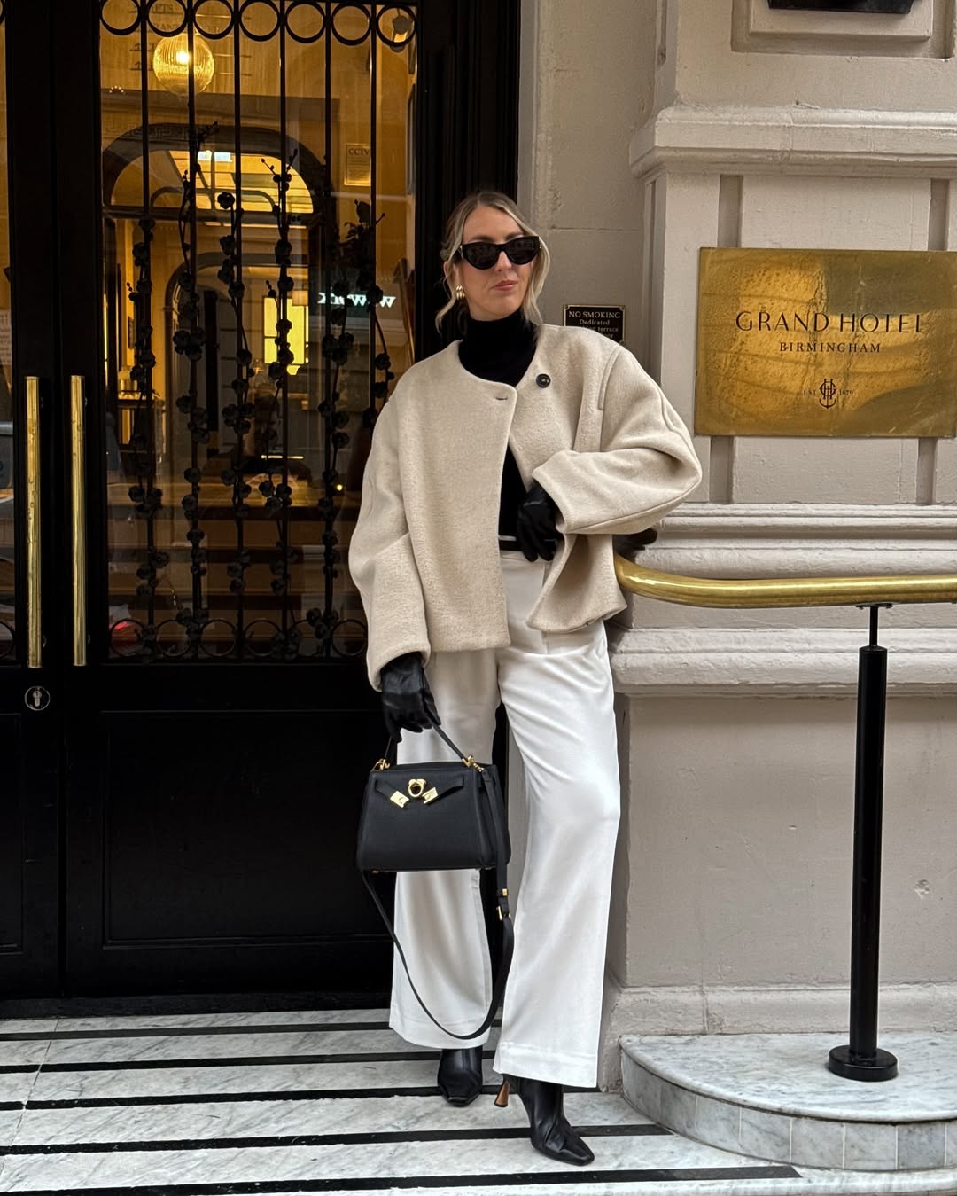 Woman in a cream textured coat with black turtleneck and white trousers.
