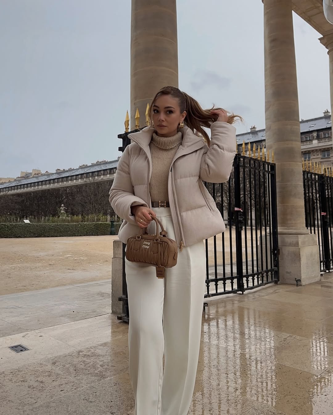 Woman in a neutral puffer coat with cream trousers and a caramel bag.