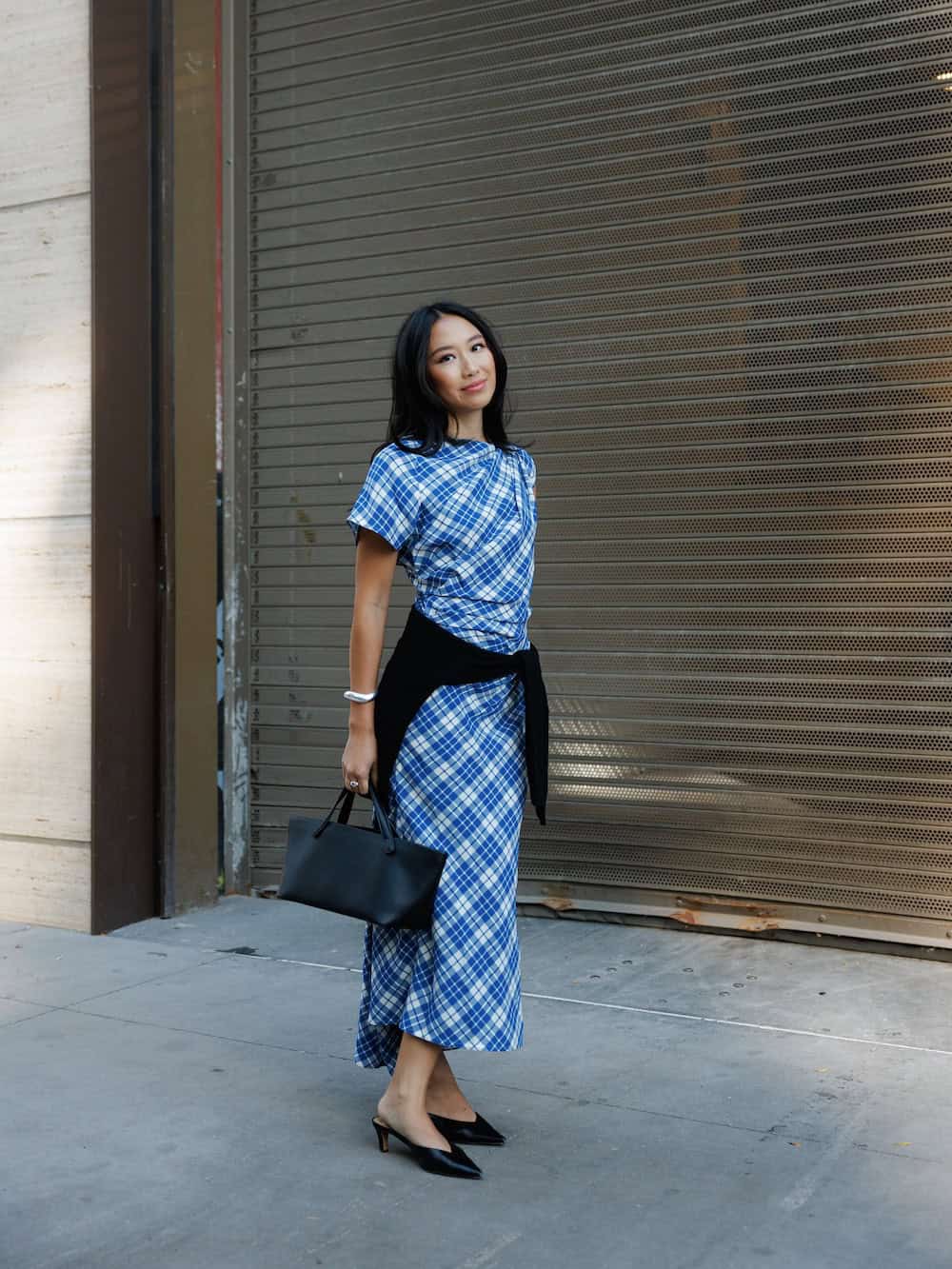 a woman wearing a blue and white plaid midi dress, a black sweater, and low black heels