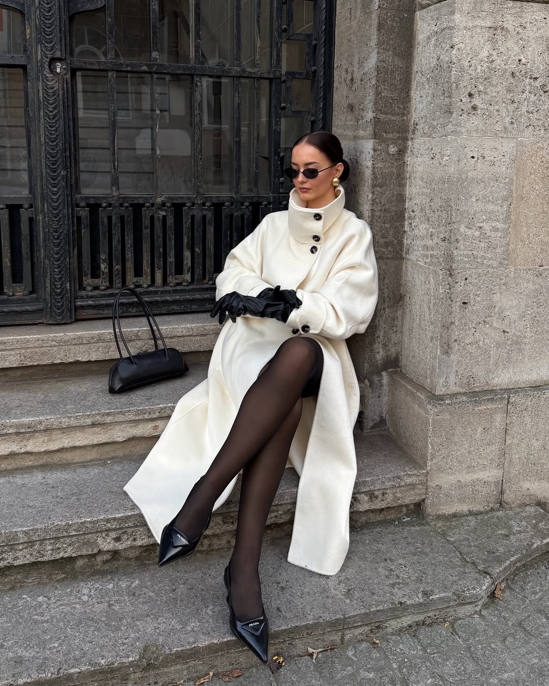 Woman wearing a long ivory coat with black tights and accessories.