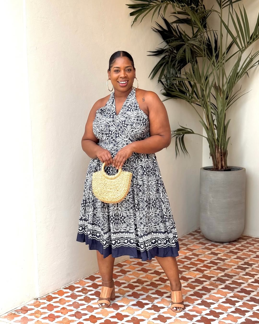 A woman in a navy and white patterned halter midi dress with a straw bag.