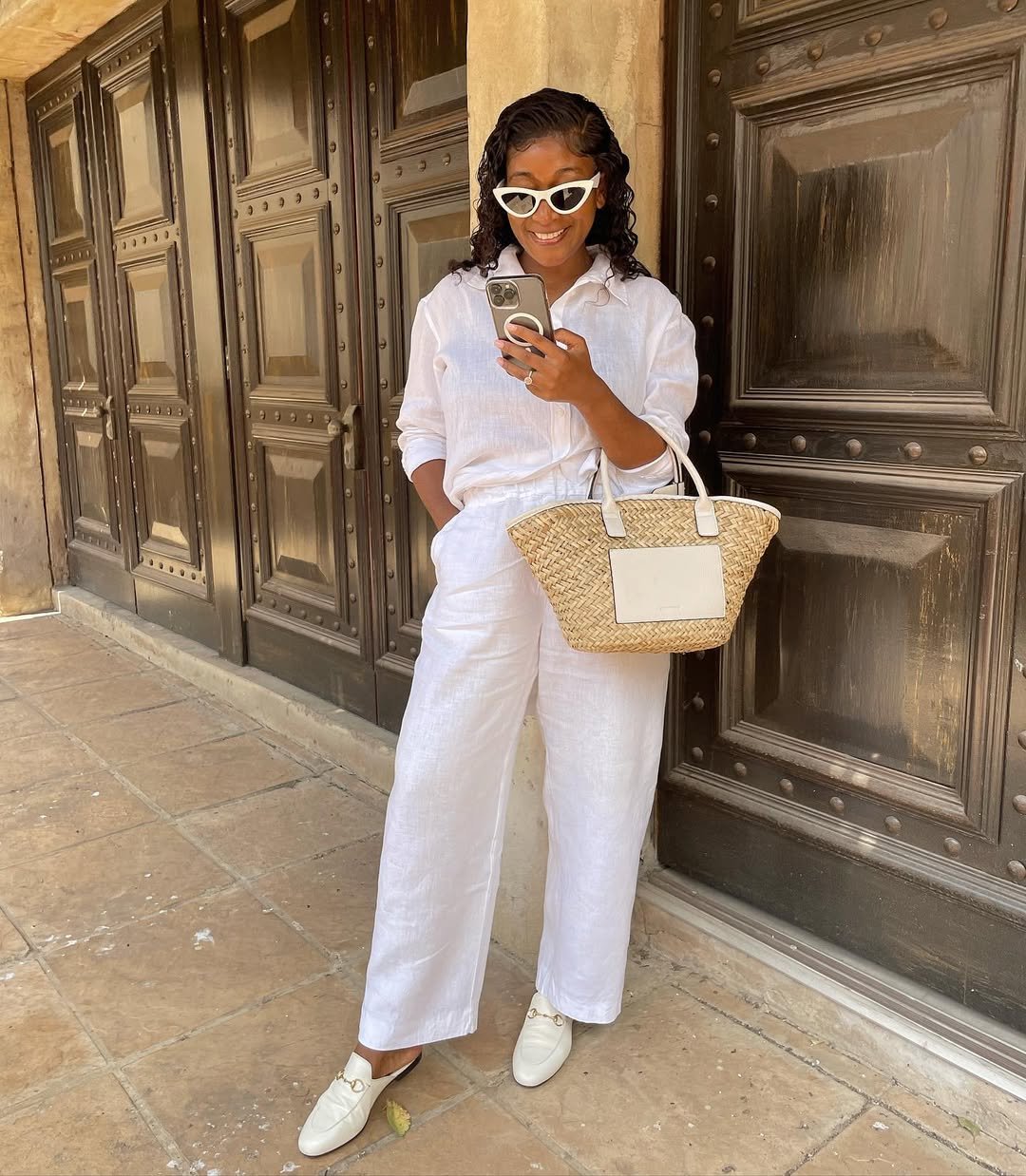Woman wearing oversized white linen shirt with matching white linen pants, a woven tote, and loafers.