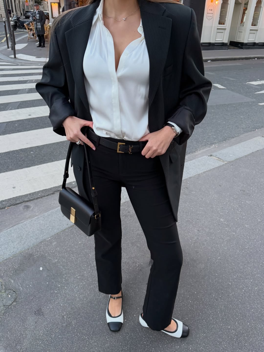 A person wears an oversized black blazer and tailored trousers with a white blouse and cap-toe flats.