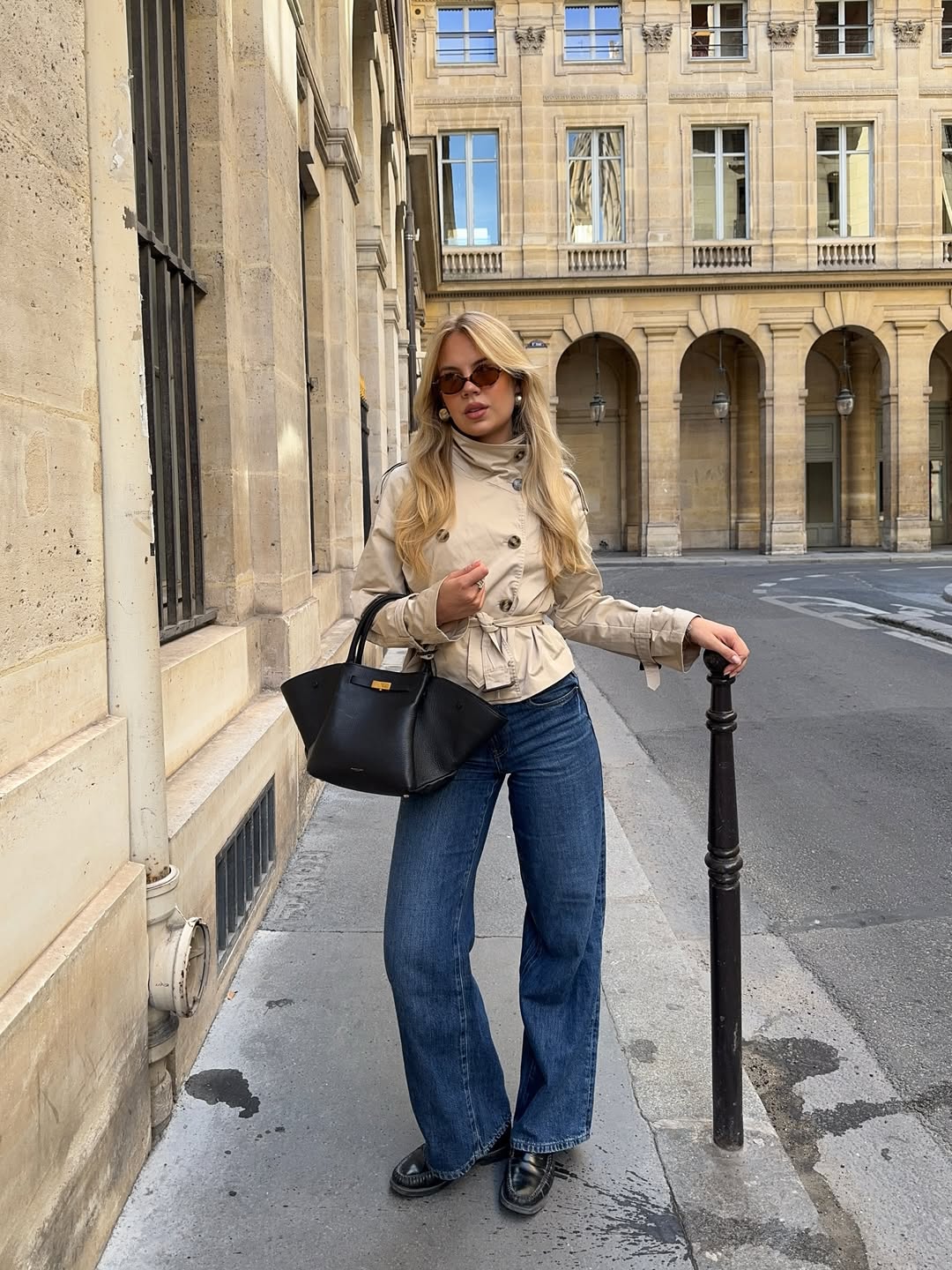 A woman wears a cropped trench jacket with wide-leg jeans, black loafers, and an oversized bag.