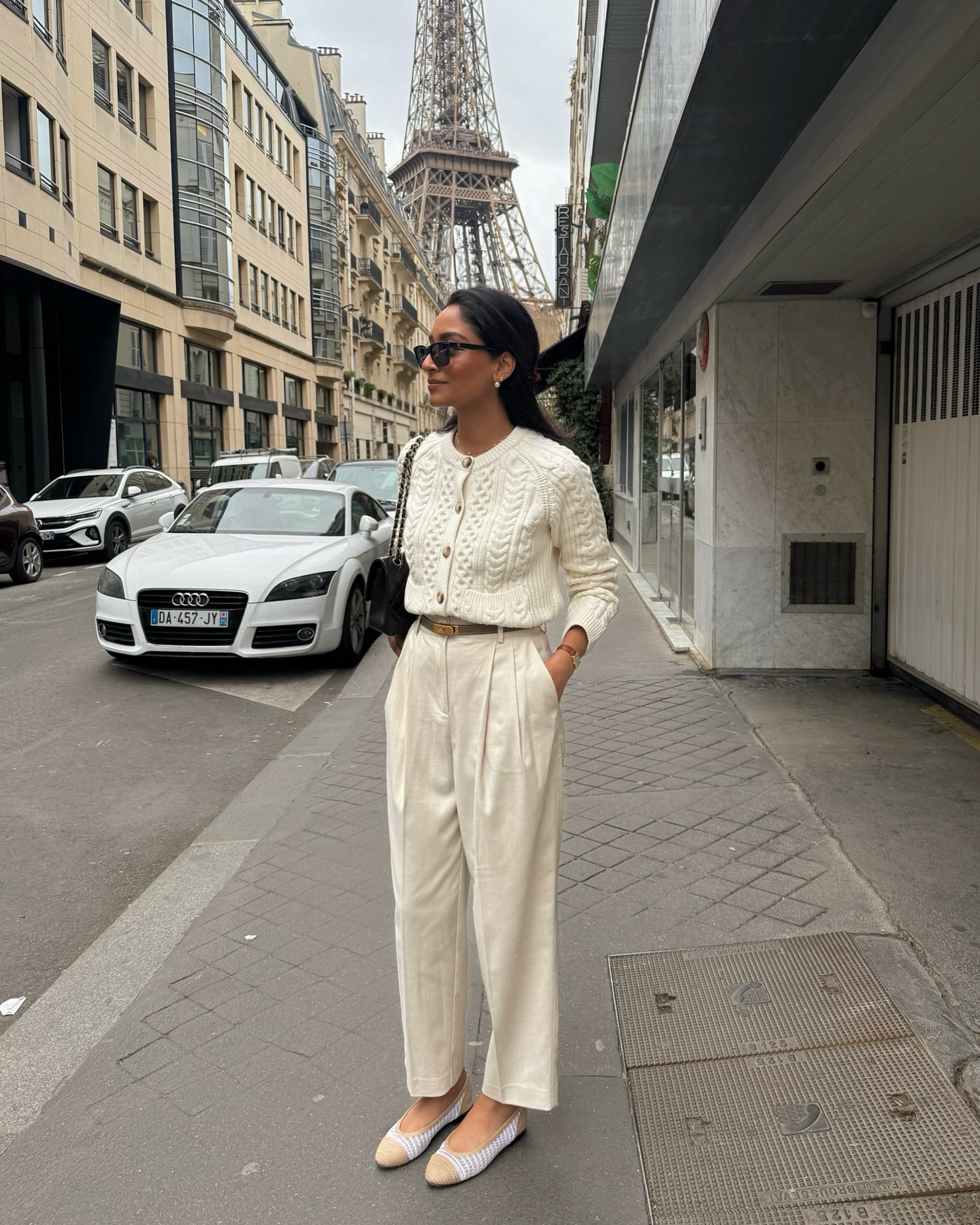 A person wears a cream cable knit cardigan with pleated trousers and woven flats.