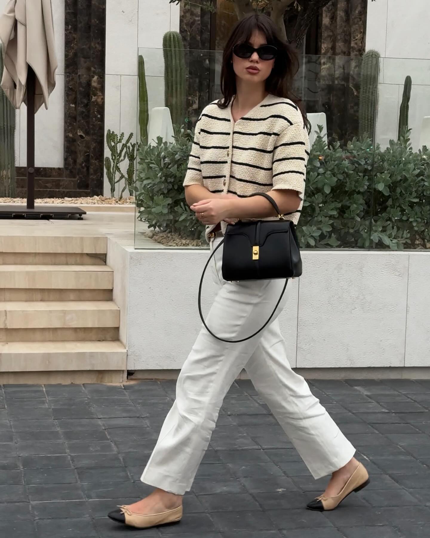 A person wears a striped knit top with white trousers, black and white flats, and a structured black bag.