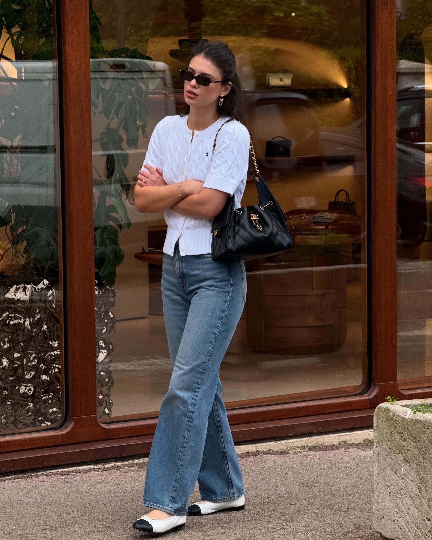 A person wears a cable knit cardigan with flared jeans, cap-toe flats, a black quilted bag, and slim sunglasses.