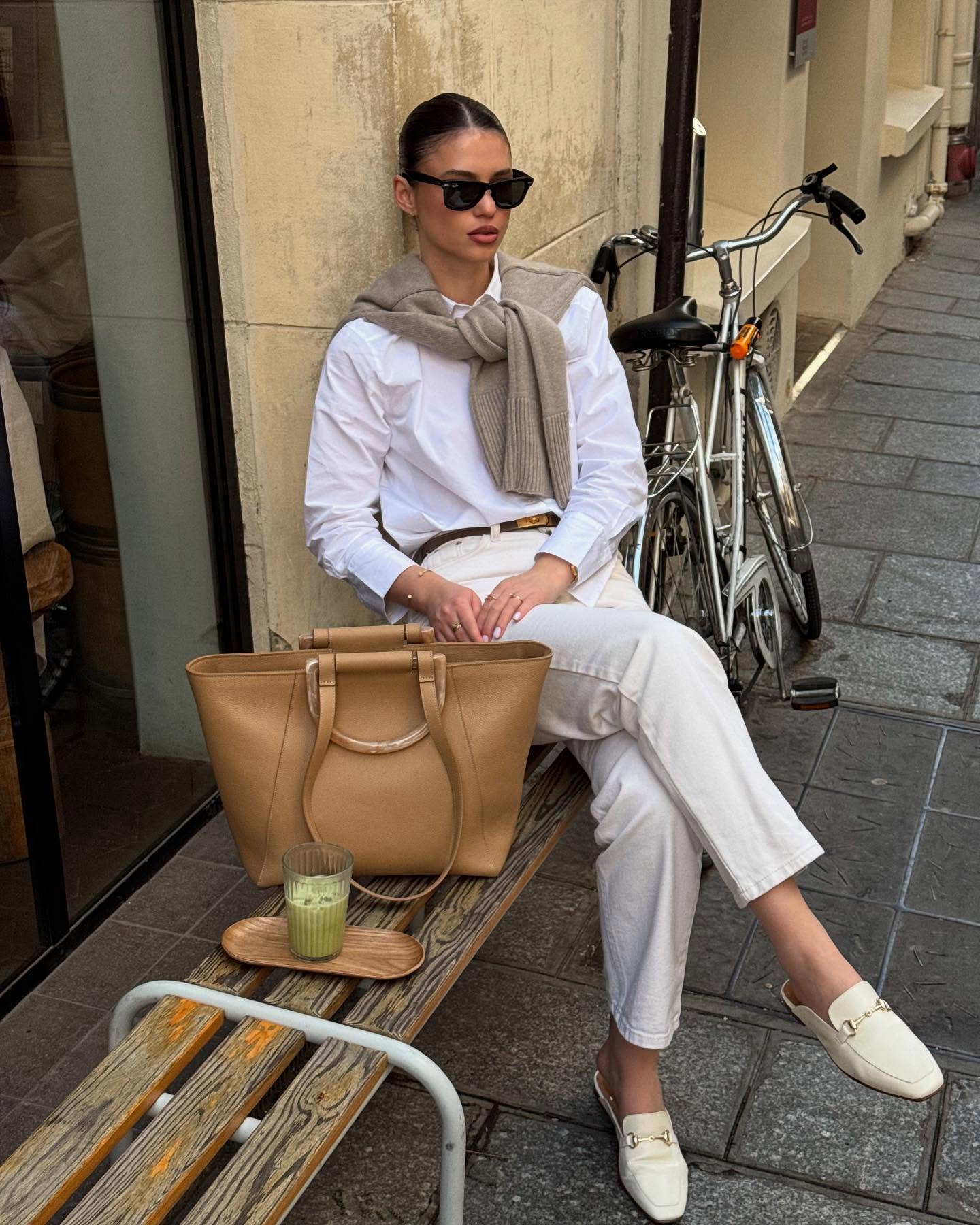 A person wears a white shirt and white jeans, with a sweater draped over their shoulders, beige tote, and loafers.