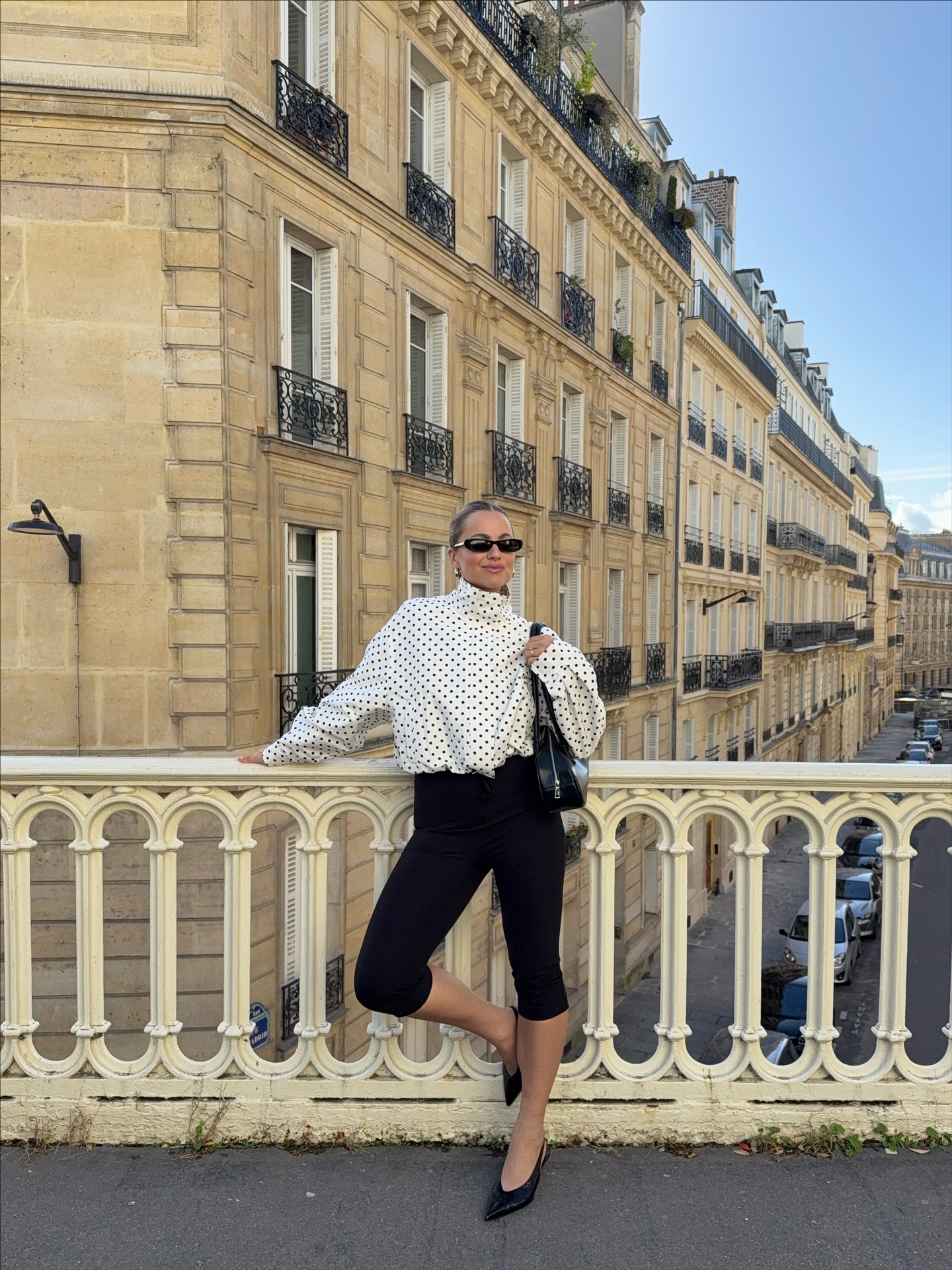 A woman wears a polka dot blouse with black capri pants, pointed heels, and a sleek bag.