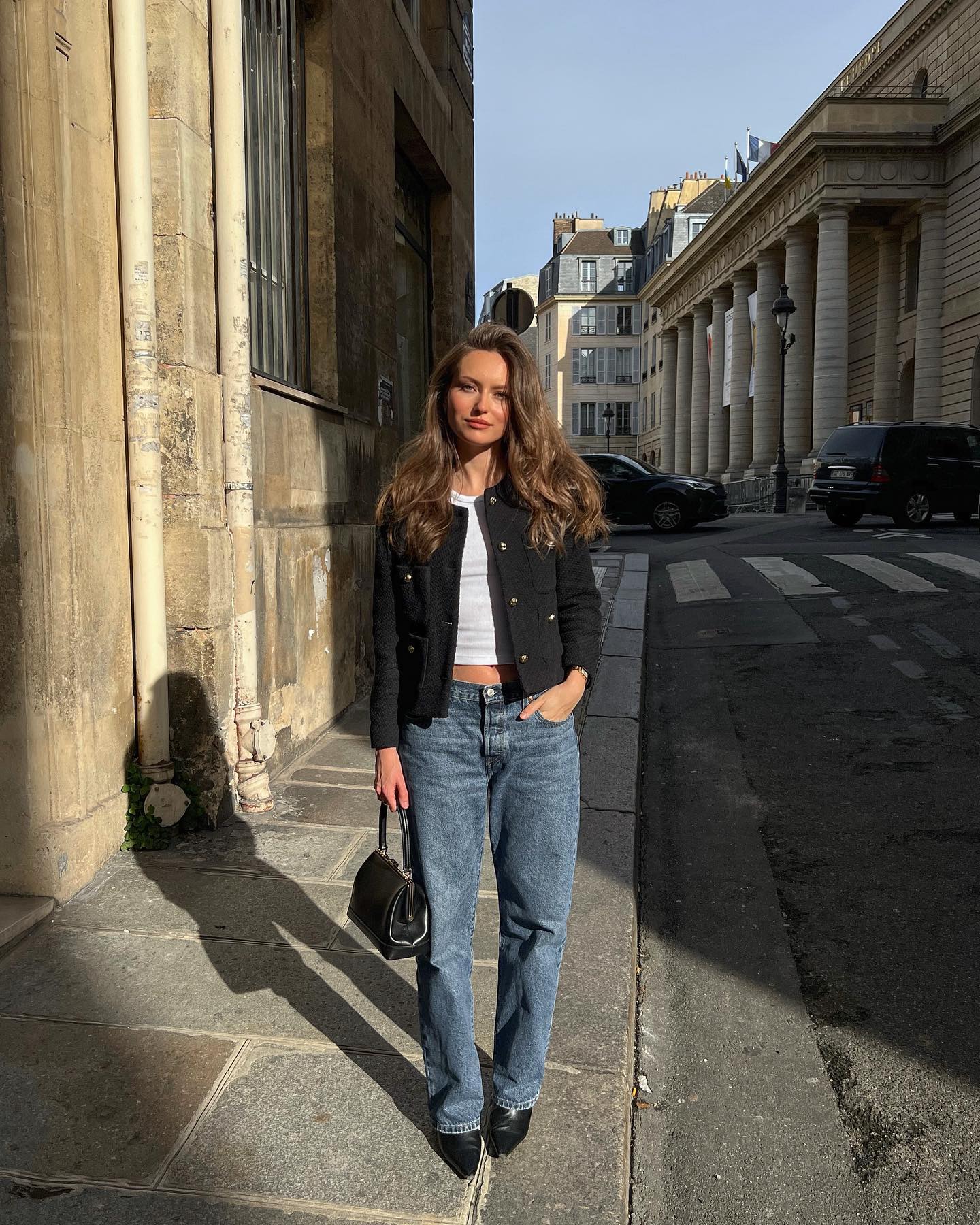 A woman wears a black cropped jacket with white top and straight-leg jeans, paired with pointed black shoes.